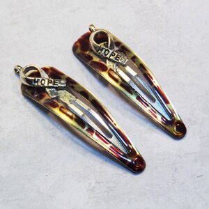 May include: Two hair barrettes with a tortoiseshell pattern in shades of brown, yellow, and red. Each barrette has a silver-colored ribbon charm with the word "HOPE" engraved on it. The barrettes are on a light gray surface.