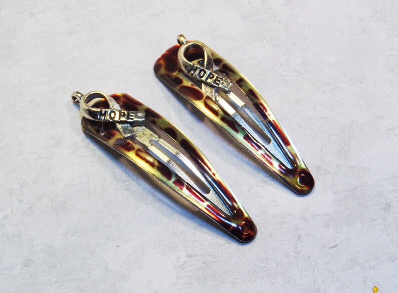 May include: Two hair barrettes with a tortoiseshell pattern in shades of brown, yellow, and red. Each barrette has a silver-colored ribbon charm with the word "HOPE" engraved on it. The barrettes are on a light gray surface.
