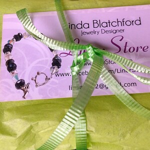 May include: A handmade bracelet with black beads and clear crystals, secured with a silver clasp, rests on a business card. The card is tied with a green ribbon and is on a green tissue paper. The card reads "Linda Blatchford Jewelry Designer".