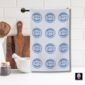 May include: A white kitchen towel with a blue and white pattern of repeated circles. Each circle contains the text "Shabbat Shalom" and a graphic of a Star of David and two candles.