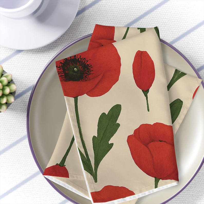 May include: Napkins with a floral poppy design. The napkins are folded and placed on a plate. The poppies are red with dark green stems and leaves. The background of the napkins is a light beige color. A white teacup and a small succulent plant are visible in the background.