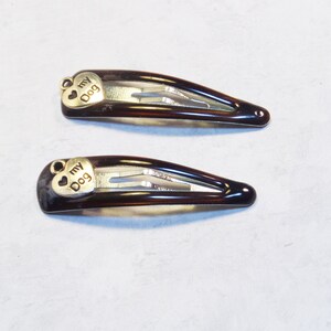 May include: Two brown hair barrettes with a tortoiseshell pattern. Each barrette has a small, gold-colored heart charm with the words "my Dog" engraved on it. The barrettes are on a light gray background.
