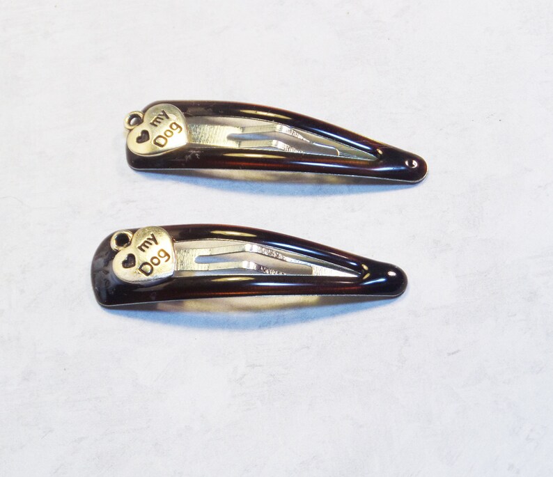 May include: Two brown hair barrettes with a tortoiseshell pattern. Each barrette has a small, gold-colored heart charm with the words "my Dog" engraved on it. The barrettes are on a light gray background.