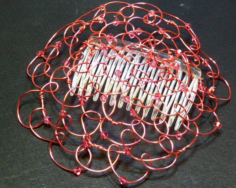 May include: A handcrafted wire kippah hair comb featuring a decorative design made of coral craft wire. The wire is intricately looped and adorned with small pink crystals. The comb itself is clear plastic, with evenly spaced teeth.