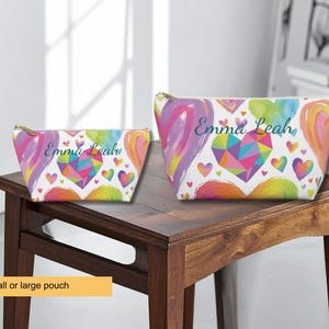 Two zippered pouches in small and large sizes, featuring a white background with a colorful pattern of watercolor hearts and a geometric heart design. The name Emma Leah is printed in a dark script font on both pouches.