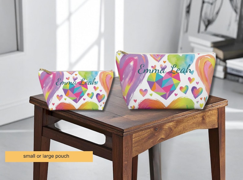 Two zippered pouches in small and large sizes, featuring a white background with a colorful pattern of watercolor hearts and a geometric heart design. The name Emma Leah is printed in a dark script font on both pouches.