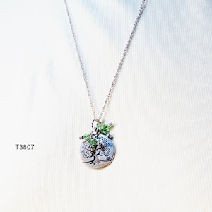 May include: A silver necklace with a circular pendant depicting a tree. The pendant is embellished with green beads. The necklace is displayed against a white backdrop. The text "T3807" is visible.
