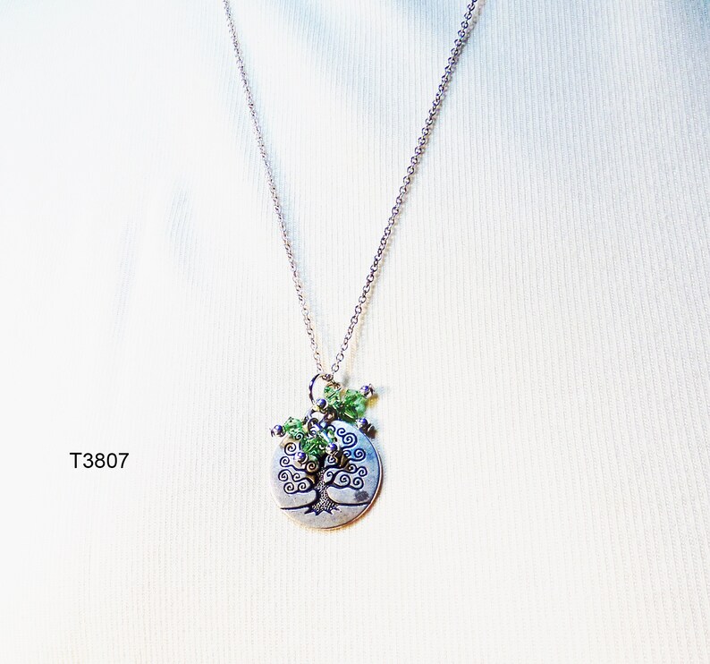 May include: A silver necklace with a circular pendant depicting a tree. The pendant is embellished with green beads. The necklace is displayed against a white backdrop. The text "T3807" is visible.