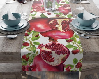 May include: A white table runner featuring a watercolor design of pomegranates, with red fruit and green leaves. The runner is set on a wooden table, with place settings, wine glasses, and a vase of pink flowers.