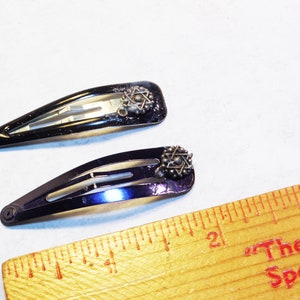 May include: Two black metal hair barrettes with a decorative starburst design. The barrettes are approximately 2 inches long. The barrettes are on a white surface.