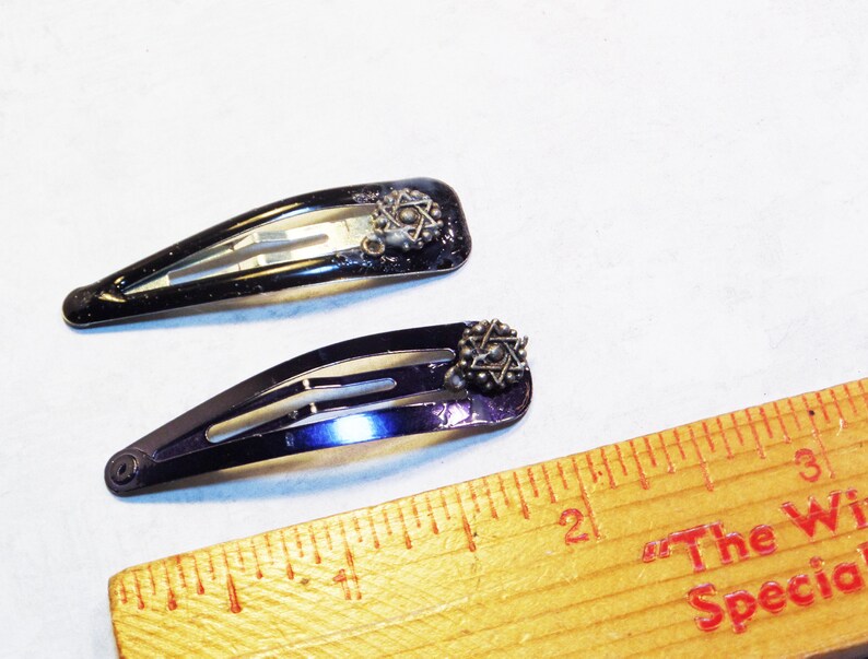 May include: Two black metal hair barrettes with a decorative starburst design. The barrettes are approximately 2 inches long. The barrettes are on a white surface.