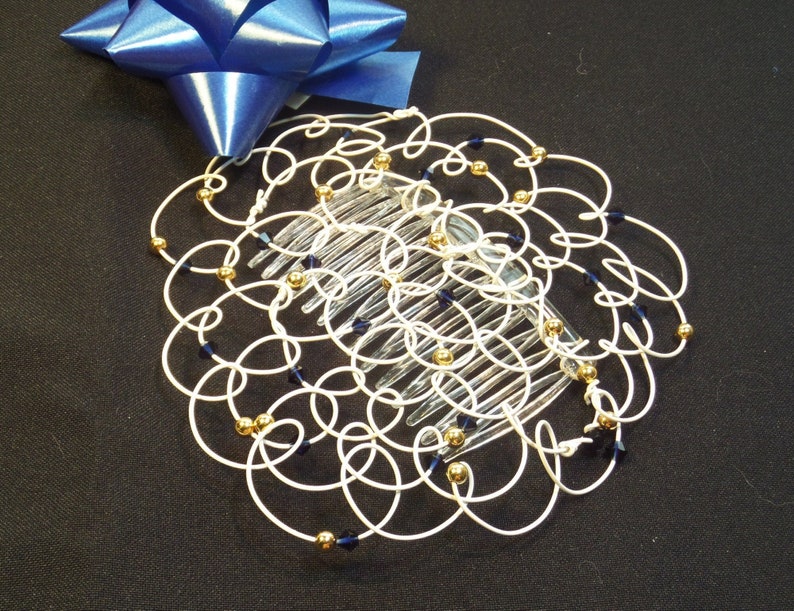 May include: A white wire hair comb with gold beads and blue crystals. The comb is in the shape of a circle and is decorated with swirls.