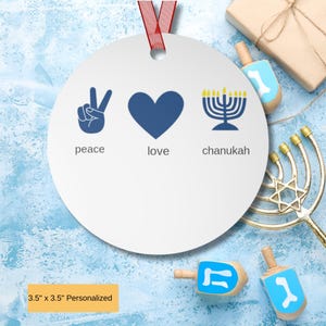May include: A round white ornament with a red ribbon featuring a blue peace sign, a blue heart, and a blue menorah with yellow flames. Below the icons, the words peace, love, and chanukah are printed in gray. The ornament measures 3.5 by 3.5 inches.