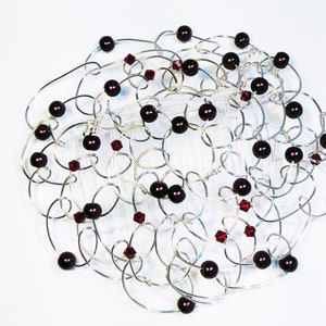 A silver-toned hair comb women's kippah with an intricate design. The comb features interconnected loops adorned with dark red beads and smaller, faceted red crystals. The design is set against a white background, highlighting the metallic and jewel tones.