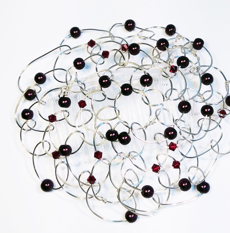 A silver-toned hair comb women's kippah with an intricate design. The comb features interconnected loops adorned with dark red beads and smaller, faceted red crystals. The design is set against a white background, highlighting the metallic and jewel tones.