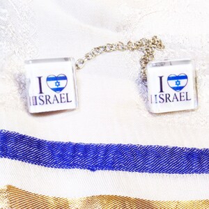 May include: Two square glass charms with a silver chain connecting them. Each charm has a white background with the text "I love Israel" and a blue and white heart with the Star of David inside.