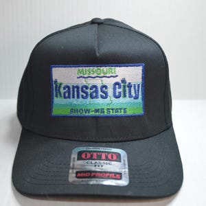 May include: Black baseball cap with a blue and green embroidered patch that says "Missouri Kansas City Show-Me State". The cap has a black bill and a black strap with a white label that says "Otto Classic Fit Mid Profile".