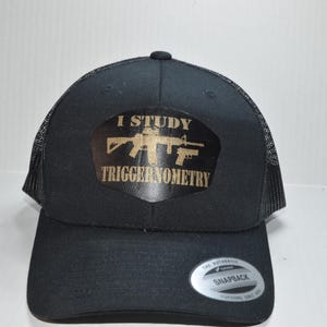 Op de afbeelding: Zwarte truckercap met een gouden patch waarop staat "Ik studeer triggernometrie" met een silhouet van een geweer.