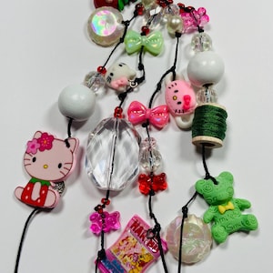 May include: A colorful beaded charm featuring various decorative elements. Includes Hello Kitty, bows, bears, and a Haribo candy package. The charm is strung on black cord and includes a red bell and a black clasp.