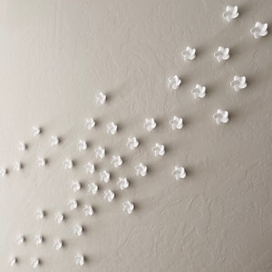 May include: White paper flowers are arranged in a diagonal pattern on a textured beige wall. The flowers are in two groups, one larger group on the left and a smaller group on the right.