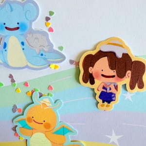 May include: A collection of colorful stickers featuring cartoon characters and heart-shaped confetti. The stickers include a blue creature, an orange dragon-like figure, and a girl with brown pigtails. The background has a pastel rainbow design.