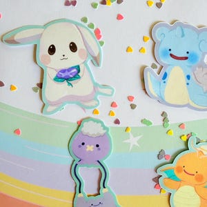 May include: A collection of colorful cartoon stickers featuring various creatures. The stickers include a white bunny holding a purple flower, a blue creature with a smiling face, and other whimsical designs. Scattered heart-shaped confetti adds a playful touch.