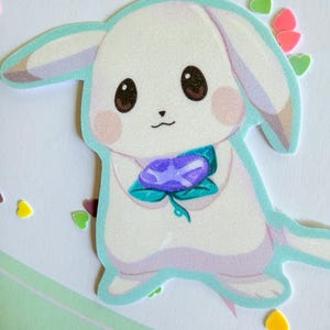 May include: A die-cut sticker of a cartoon bunny with a white body, brown eyes, and pink cheeks. The bunny holds a purple flower with green leaves. The sticker has a light blue outline and is surrounded by heart-shaped confetti.