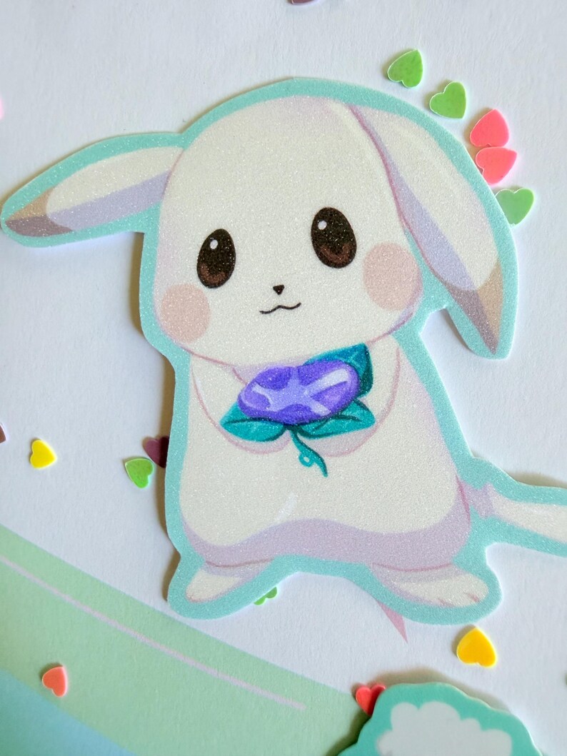 May include: A die-cut sticker of a cartoon bunny with a white body, brown eyes, and pink cheeks. The bunny holds a purple flower with green leaves. The sticker has a light blue outline and is surrounded by heart-shaped confetti.