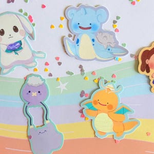 May include: A collection of colorful cartoon stickers featuring various cute characters. The stickers include a white bunny with a flower, a blue sea creature, a girl in a dress, a purple creature, and an orange dragon. The background is a pastel rainbow.