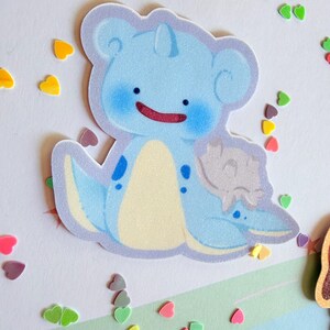 May include: A light blue cartoon sticker of a creature with a smiling red mouth, blue cheeks, and a cream-colored belly. The sticker is surrounded by colorful heart-shaped confetti.