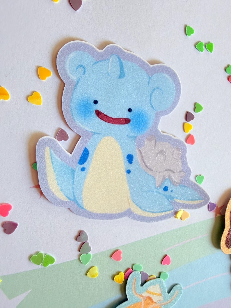 May include: A light blue cartoon sticker of a creature with a smiling red mouth, blue cheeks, and a cream-colored belly. The sticker is surrounded by colorful heart-shaped confetti.