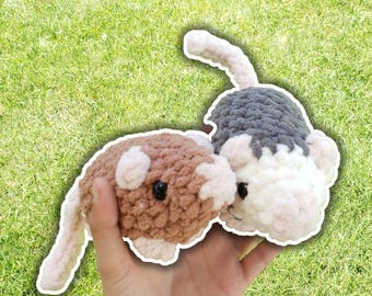 May include: Two handmade crochet mice in shades of brown, white, and gray. The mice have small black eyes and long tails. The toys are held against a backdrop of green grass.