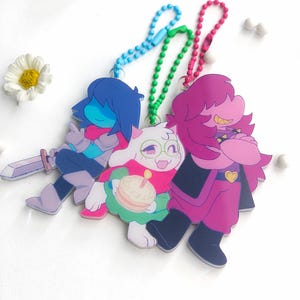 May include: Three acrylic character charms with ball chains. One charm features a blue-haired figure with a sword, another shows a white creature holding a cake, and the third depicts a pink-haired figure with a yellow heart emblem on their chest.
