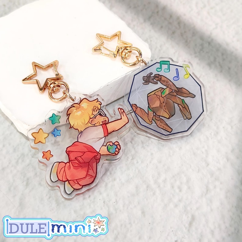 May include: Two acrylic keychains with gold-colored star-shaped clasps. One keychain features a cartoon character with a red shirt and pants, reaching for colorful stars. The other keychain has a geometric design with a brown figure and musical notes.