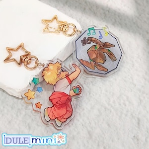 May include: Clear acrylic keychains with gold-colored hardware. One keychain features a cartoon figure in red pants and a white shirt, reaching for a ball. Another keychain has a geometric design with a brown figure and musical notes. The brand name "DULE mini" is at the bottom.