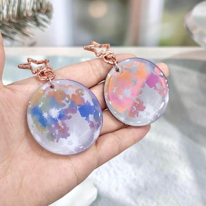 May include: Two circular keyrings with a celestial design. Each keyring features a colourful moon graphic with shades of blue, orange, and pink. The keyrings have a rose gold star-shaped clasp.