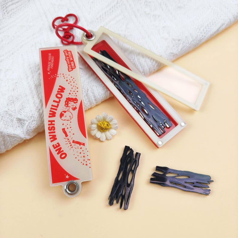 Op de afbeelding: Een rechthoekige, doorschijnende houder met rood-witte afbeeldingen met de tekst ONE WISH WILLOW, AMAZE YOUR FRIENDS en You only get ONE WISH. De houder bevat donkere, poreuze, takachtige stukjes, met twee extra stukjes en een kleine witte bloem.