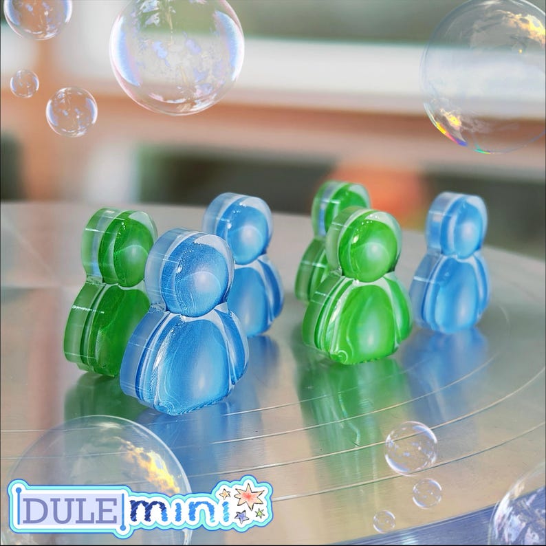 K&ouml;nnte beinhalten: Eine Sammlung transluzenter, stilisierter Figuren in Form von Menschen in Blau- und Gr&uuml;nt&ouml;nen. Die Figuren sind auf einer reflektierenden Oberfl&auml;che angeordnet, mit Blasen im Hintergrund. Das Logo "DULE mini" ist unten sichtbar.
