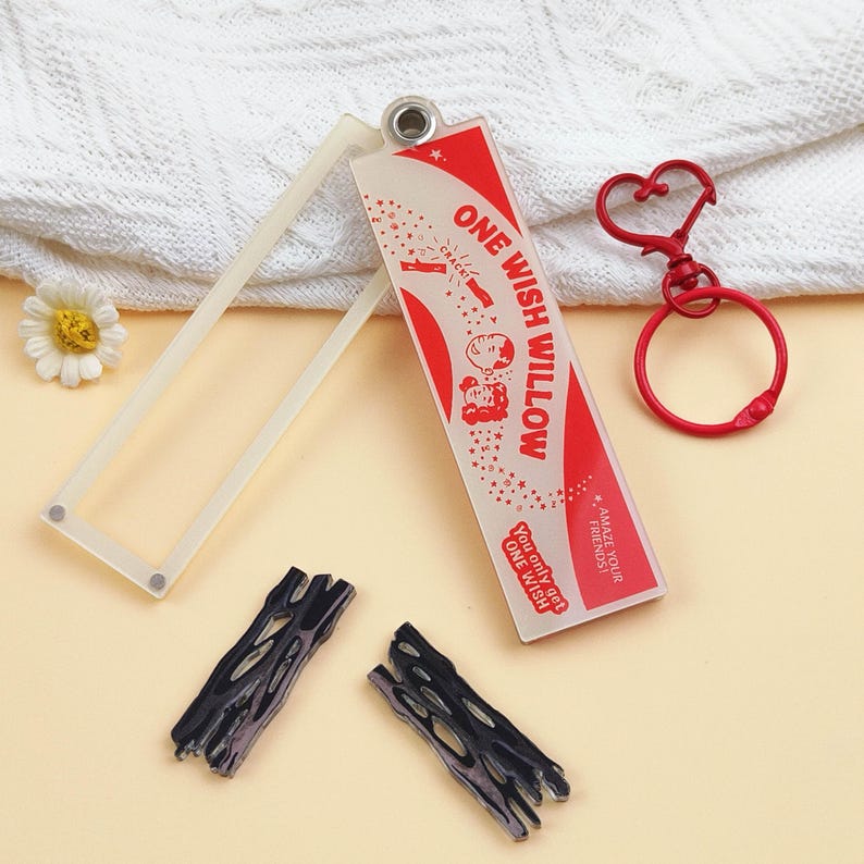 Op de afbeelding: Een rechthoekige acryl sleutelhangercomponent met een rood en wit ontwerp met de tekst ONE WISH WILLOW, You only get ONE WISH en AMAZE YOUR FRIENDS. Het bevat een bijpassende rode hartvormige sluiting, een transparant frame en twee donkere, getextureerde, takvormige bedels.
