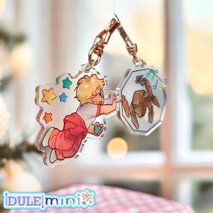 May include: Two clear acrylic keychains with gold-colored hardware. One keychain features a cartoon boy in red overalls and a white shirt, the other a brown creature with musical notes. The brand name "DULE mini" is at the bottom.