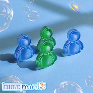 K&ouml;nnte beinhalten: Drei durchscheinende Figuren in Blau- und Gr&uuml;nt&ouml;nen sind auf einer hellblauen Oberfl&auml;che angeordnet. Die Figuren haben eine abgerundete, abstrakte menschen&auml;hnliche Form. Blasen schweben im Hintergrund, und der Text "DULE mini" ist unten sichtbar.
