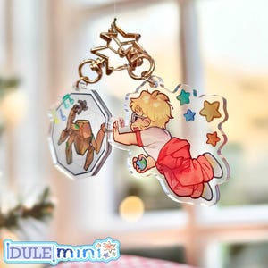 May include: Clear acrylic charm featuring a cartoon illustration of a person reaching towards a geometric shape with a robot inside. The charm has a gold star-shaped clasp and a smaller charm with a robot illustration. The person is wearing glasses, a white shirt, and red pants.