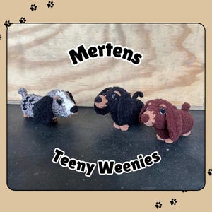 May include: Three crocheted dachshund dog plushies. The plushies are black and white, black, and brown. The plushies are sitting on a wooden surface. The text "Mertens Teeny Weenies" is written on the surface.