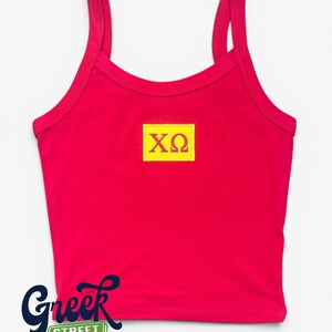 May include: A red crop top with spaghetti straps. A yellow rectangle with the Greek letters ΧΩ is centered on the front. The words "Greek Street" are in a navy blue script font at the bottom, with the words "Greek Life" and "Styled Right" below.