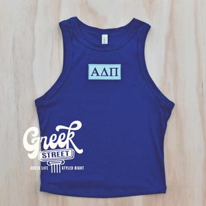 May include: A blue tank top with the Greek letters ΑΔΠ on a light blue rectangle. The tank top has a scoop neck and is sleeveless. The words "Greek Street" are printed in white script on the bottom left.