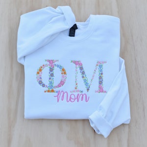 May include: White sweatshirt with the letters "OM" embroidered in colorful flowers. The word "mom" is embroidered in pink below the letters.