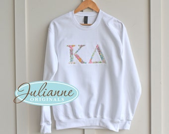 May include: White sweatshirt with a floral embroidered "KA" design.