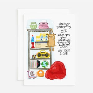 May include: Greeting card with a whimsical illustration of a shelf filled with vintage items, including a typewriter, lava lamp, and boombox. The card features the text: "You know you're getting OLD when you find treasures from your childhood in an ANTIQUE STORE!"