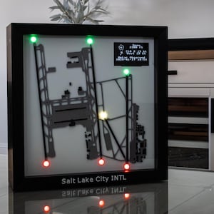 Könnte beinhalten: Ein gerahmtes, beleuchtetes Flughafendiagramm mit schwarzem Rahmen. Das Diagramm zeigt ein graues Layout von Start- und Landebahnen und Rollwegen, mit grünen und roten LED-Leuchten. Der Text "Salt Lake City INTL" ist unten zusammen mit Wetterdaten zu sehen.