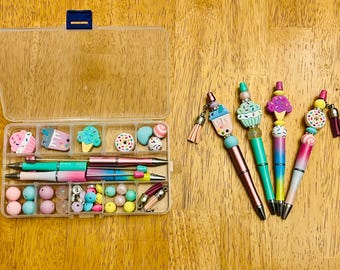 Op de afbeelding: Een doorzichtige plastic organizerbox met een blauwe sluiting, gevuld met kleurrijke kralen, bedels en pennen. De pennen hebben decoratieve toppen in de vorm van cupcakes, ijsjes en donuts. De pennen zijn roze, blauw en groen.